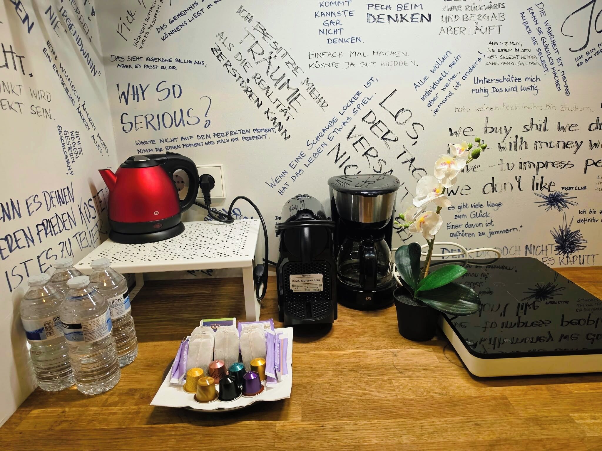 Ein Zimmer mit einem Holztisch, einem roten Wasserkocher, einer Kaffeemaschine, einer Pflanze und Wasserflaschen.