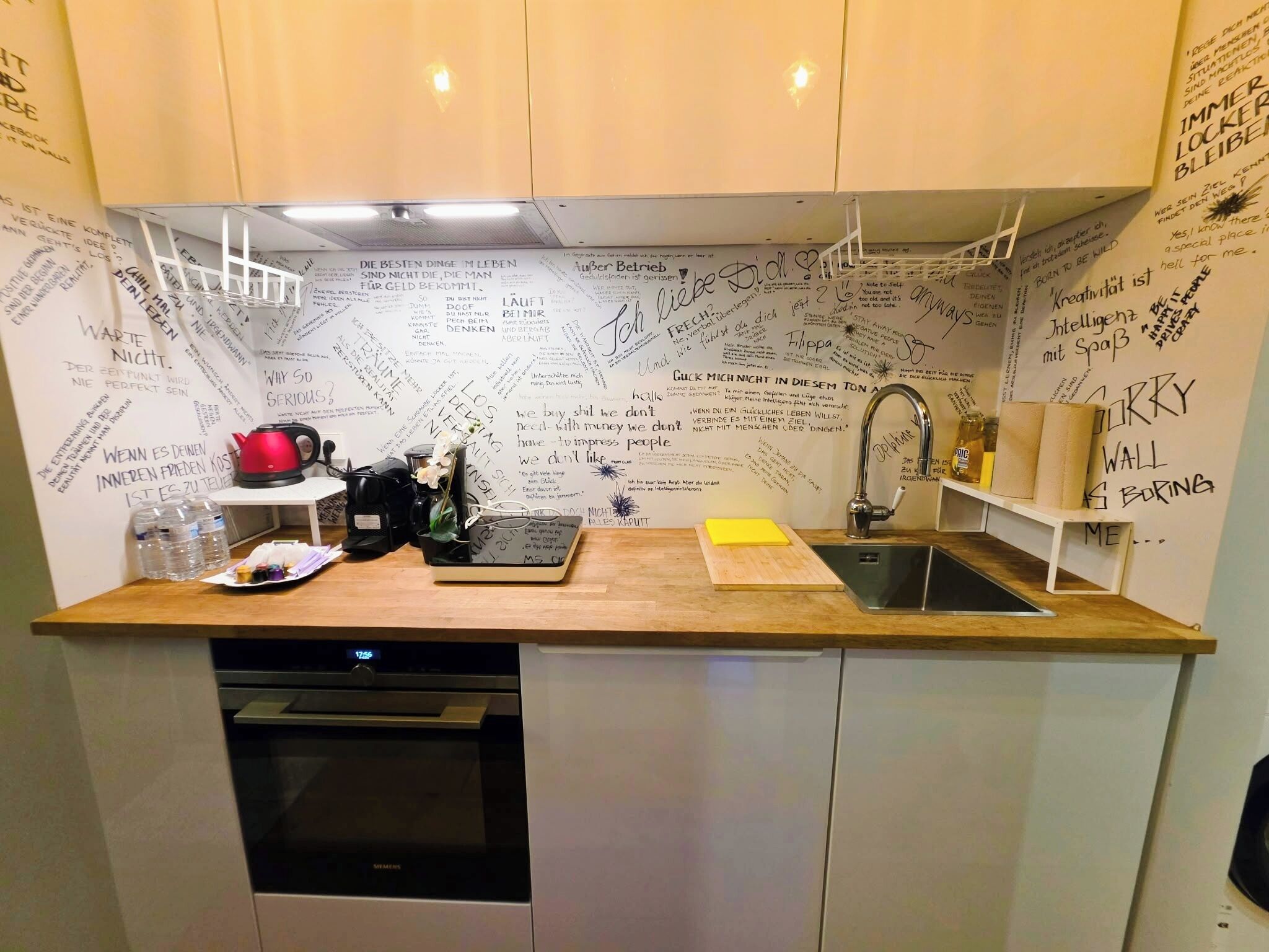 Een kleine keuken met een houten aanrecht, een roestvrijstalen spoelbak en een vaatwasser. De muren zijn bezaaid met handgeschreven aantekeningen en tekeningen.