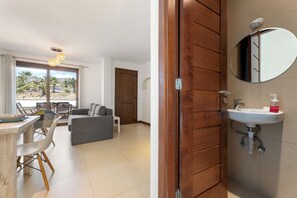 Una habitación con comedor, sofá, puerta de madera y baño con lavamanos y espejo.