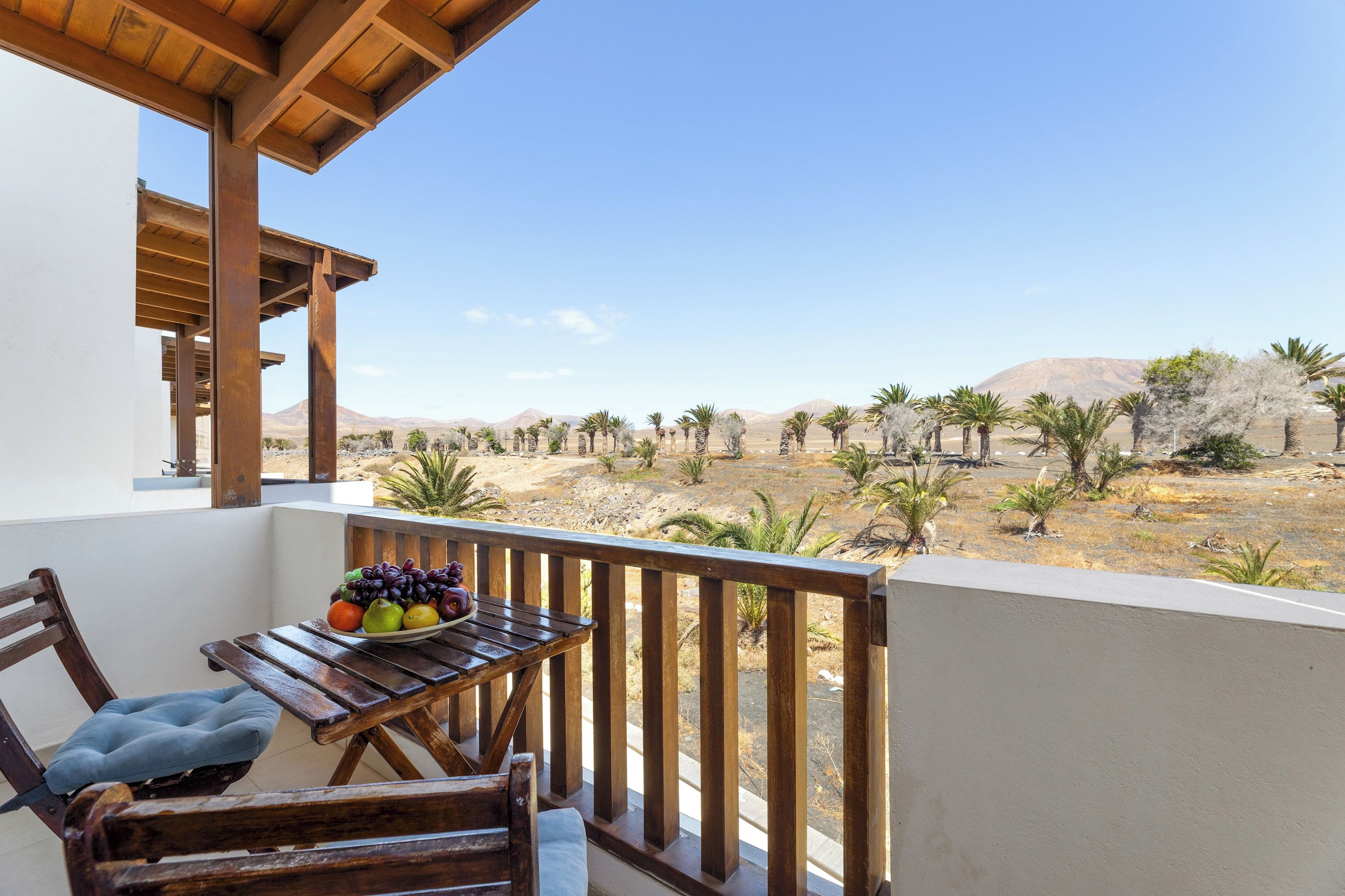 A balcony with a wooden table and chairs, overlooking a desert landscape with sparse vegetation and distant mountains.