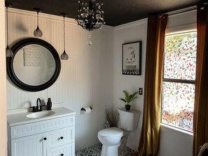 Ein Badezimmer mit weißem Waschtisch, einem runden Spiegel, einer Kristallleuchtre, einer Toilette und einem Fenster mit Vorhängen.