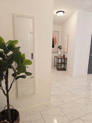 Un pasillo con una planta en maceta, una puerta, piso de baldosas y una mesita con un florero.