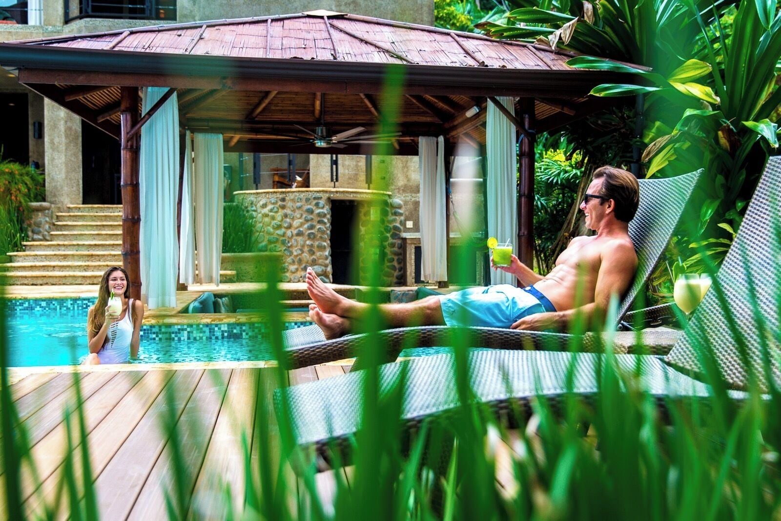 A pool area with a lounge chair, a person relaxing, and a woman in the water.