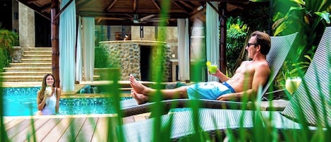Un área de piscina con una silla de descanso, una persona relajándose y una mujer en el agua.