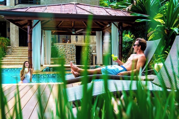A pool area with a lounge chair, a person relaxing, and a woman in the water.