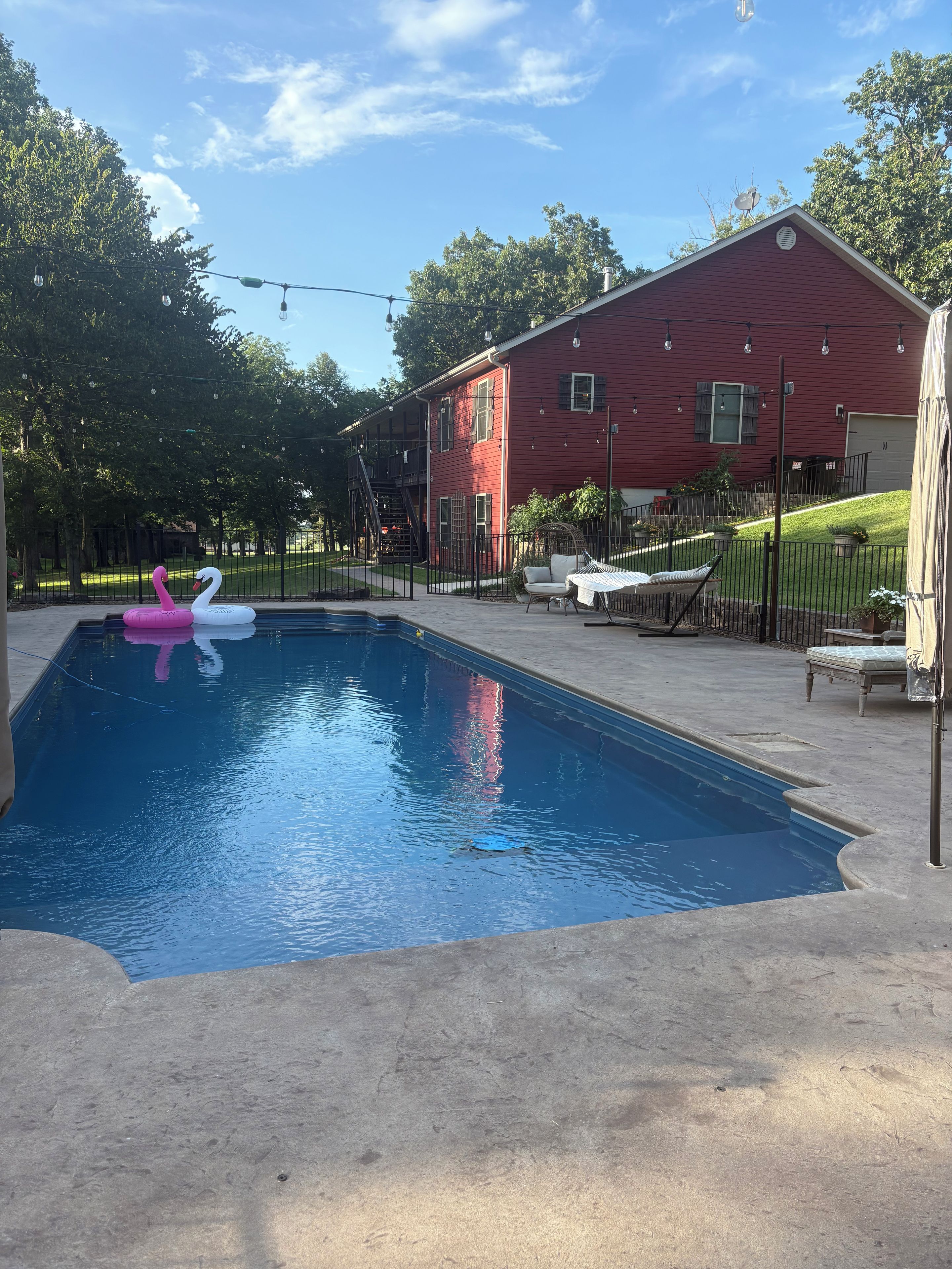 A swimming pool with a red building in the background, a fenced yard, and outdoor furniture.