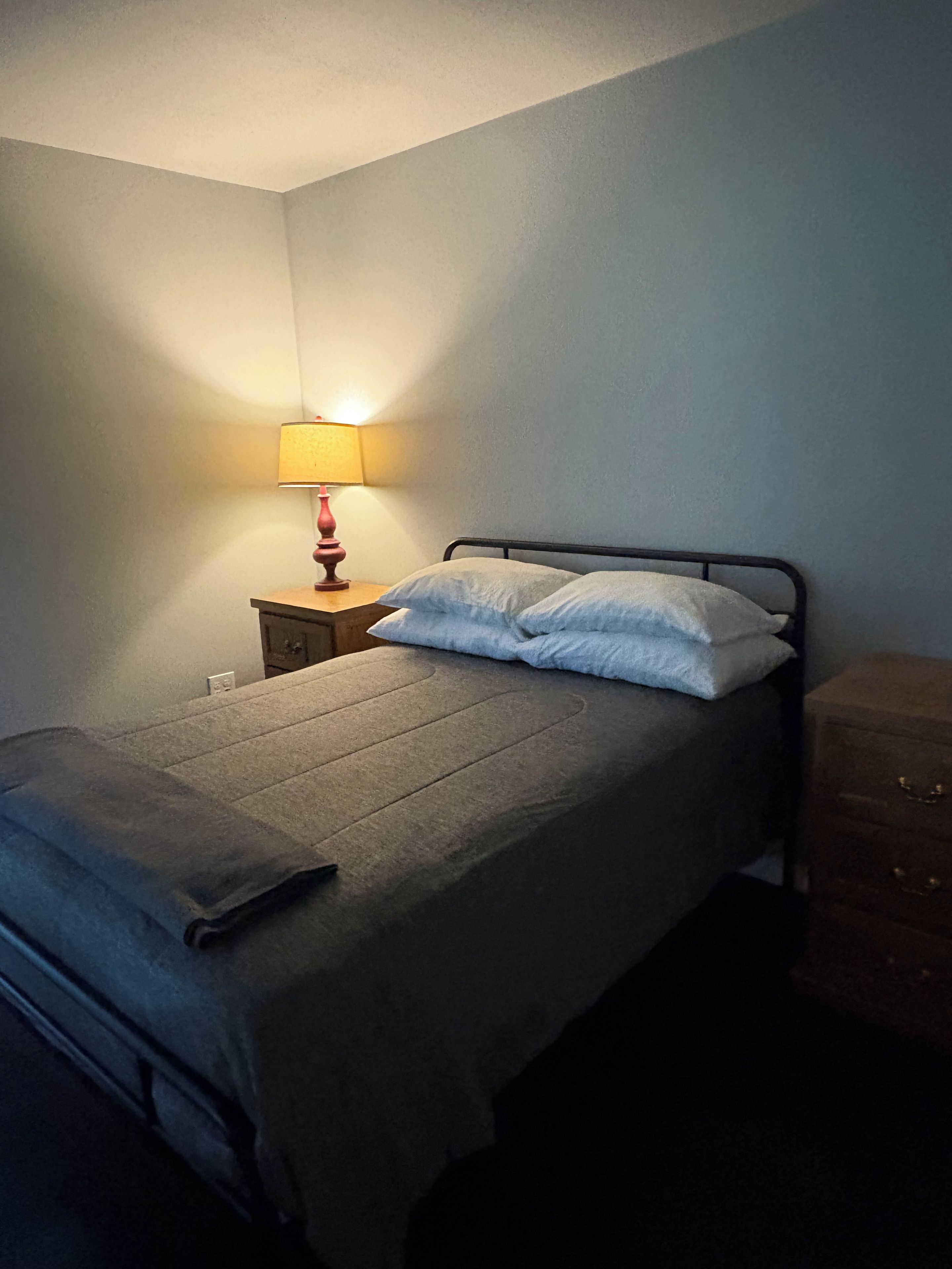 A single bed with a grey bedspread and two pillows, a bedside table with a lamp, and a wooden dresser in a room with plain walls.