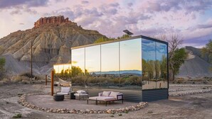 A modern outdoor lounge with a glass enclosure, furnished with a sofa and chairs, set against a desert landscape with a mountain in the background.
