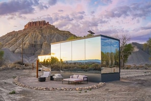 A modern outdoor lounge with a glass enclosure, furnished with a sofa and chairs, set against a desert landscape with a mountain in the background.