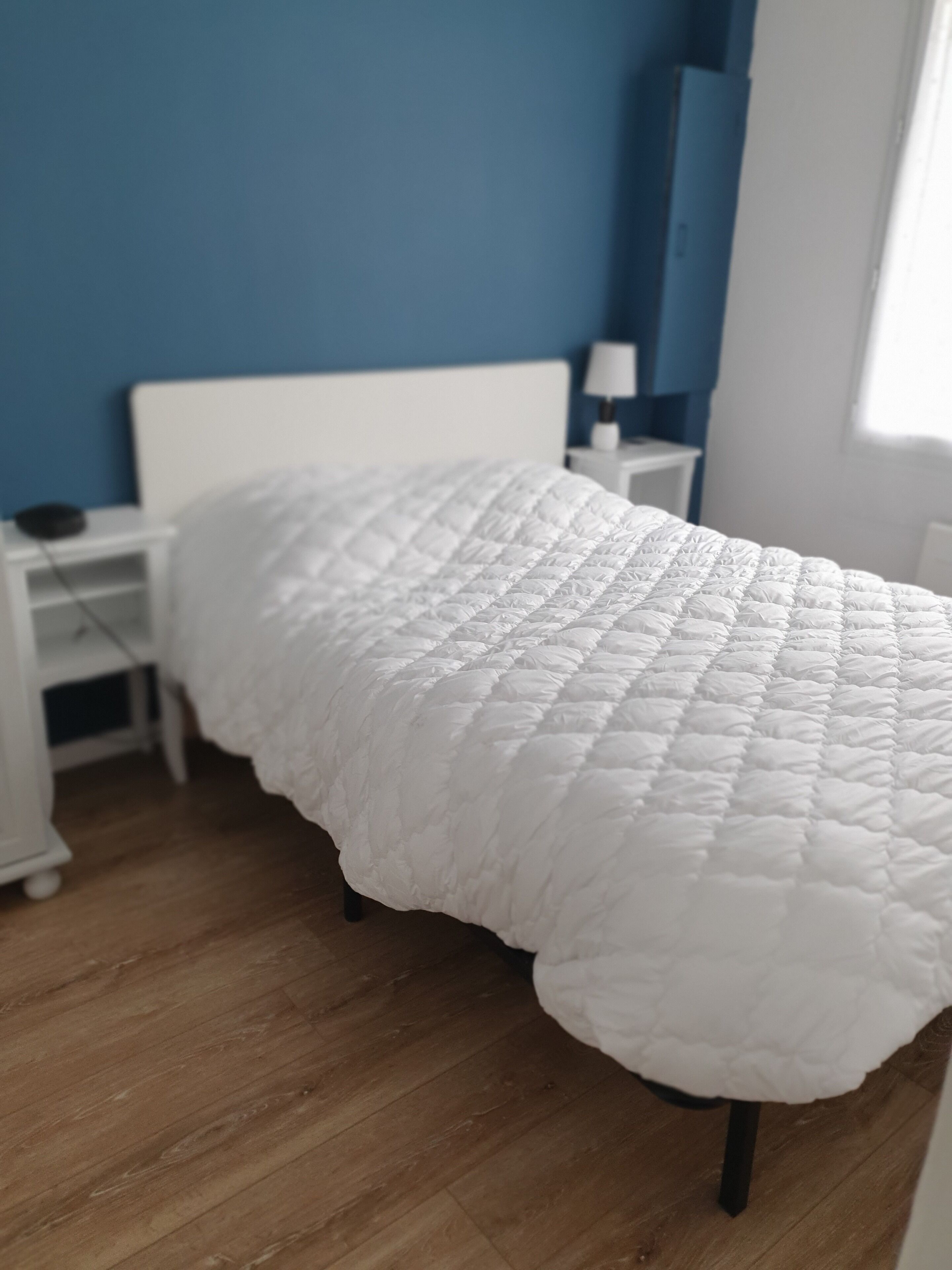 Uma cama com colcha branca em quarto com paredes azuis e chão de madeira.