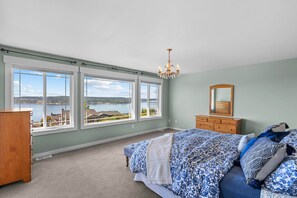 A bedroom with a large bed, a dresser, a mirror, and a chandelier. There are three large windows providing a view of a lake and buildings. - Chamber's Bay Lodging (University Place)
