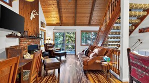 A spacious living room with a fireplace, wooden flooring, a leather sofa, and a staircase with patterned carpet. - Wilderness Lodge by Avantstay Firepit Gazebo + Wraparound Decks Minutes to Village + Trails (Cedar Glen)