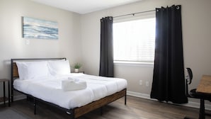 Quarto de hotel com cama, escrivaninha, cadeira, janela com cortinas, ventilador de teto e um quadro na parede.