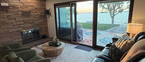 A living room with a stone wall, a fireplace, a sofa, and a view of a body of water through large sliding doors.