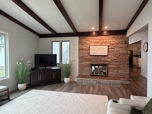 A living room with a stone fireplace, a television, and a view of the kitchen.