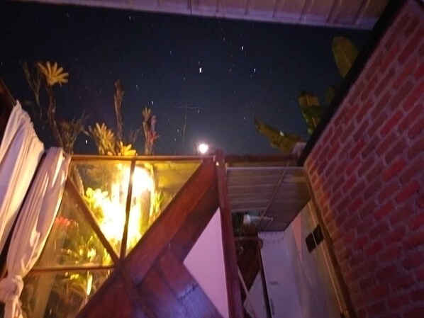 Escena nocturna con cielo estrellado, una ventana iluminada y un muro de ladrillo.