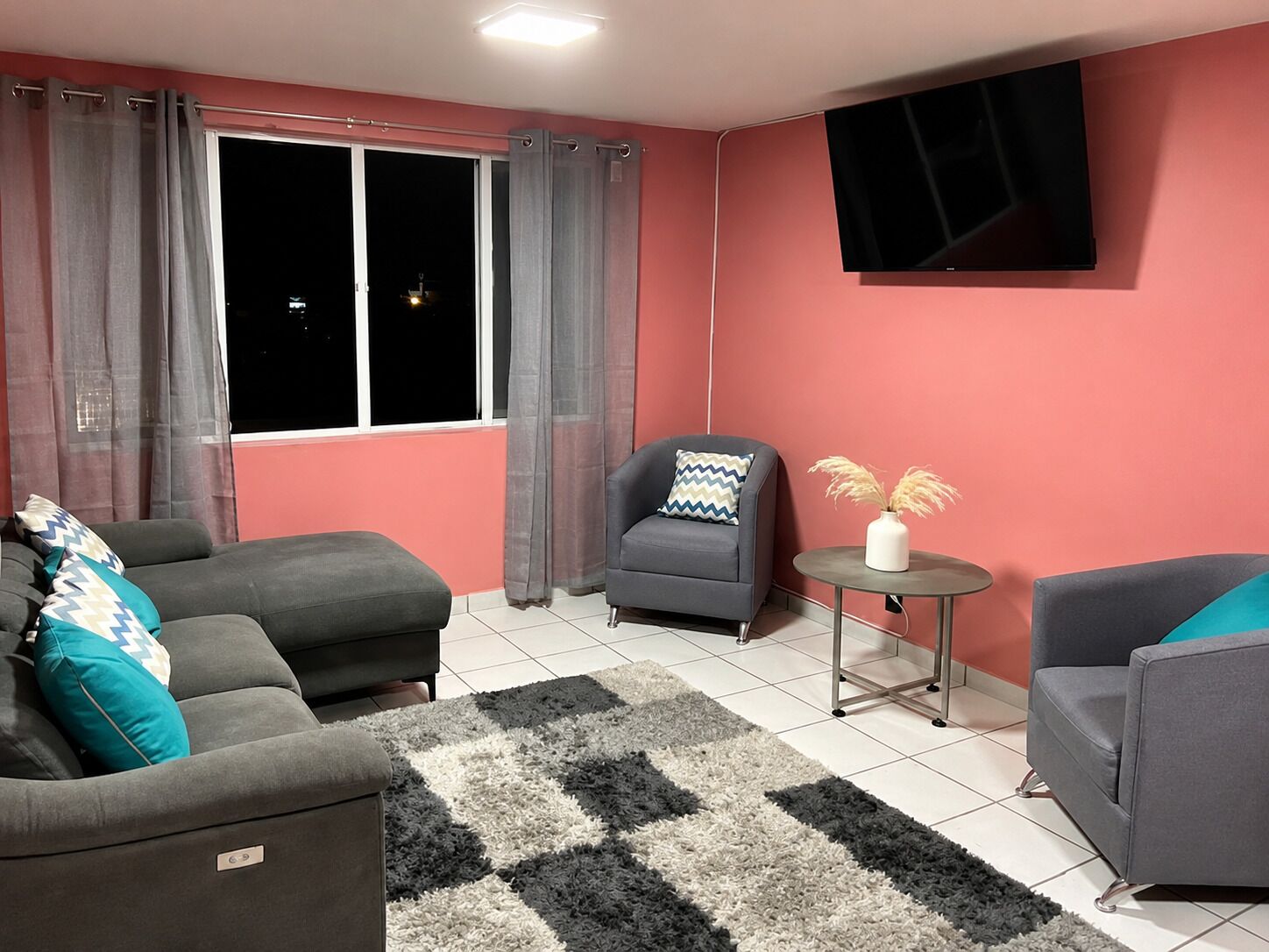 Una sala de estar con un sofá, un sillón, una mesa redonda con un florero y un televisor montado en la pared.