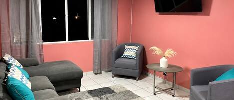 Una sala de estar con un sofá, un sillón, una mesa redonda con un florero y un televisor montado en la pared.