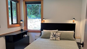 Ein Schlafzimmer mit Bett, Nachttischlampen, einem Nachttisch, einem Schreibtisch und einem Fenster mit Blick auf Grünflächen.