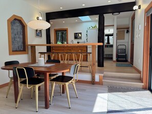 Un comedor con mesa y sillas de madera, una lámpara colgante y vista a la cocina a través de un vano abierto.