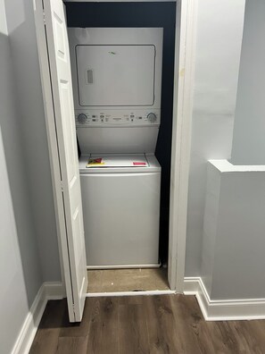 A laundry room with a front-loading washer and dryer, a control panel, and a small shelf with items.