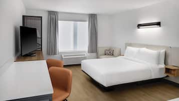 Ein Hotelzimmer mit Bett, Schreibtisch, Stuhl, Fernseher, einem Fenster mit Vorhängen und einer Wandleuchte.