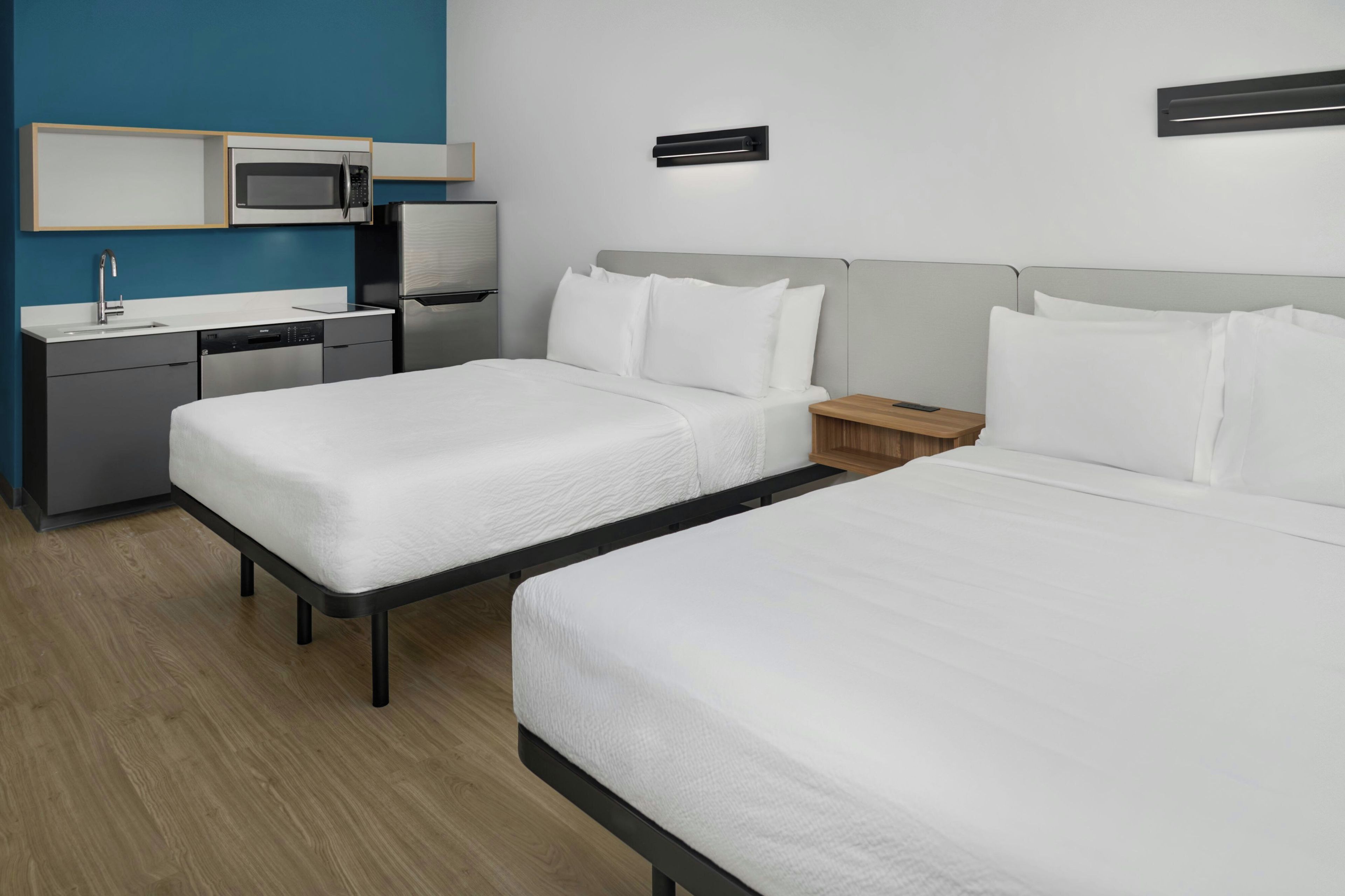 Et hotelværelse med to senge, et lille tekøkken og et natbord med en lampe.