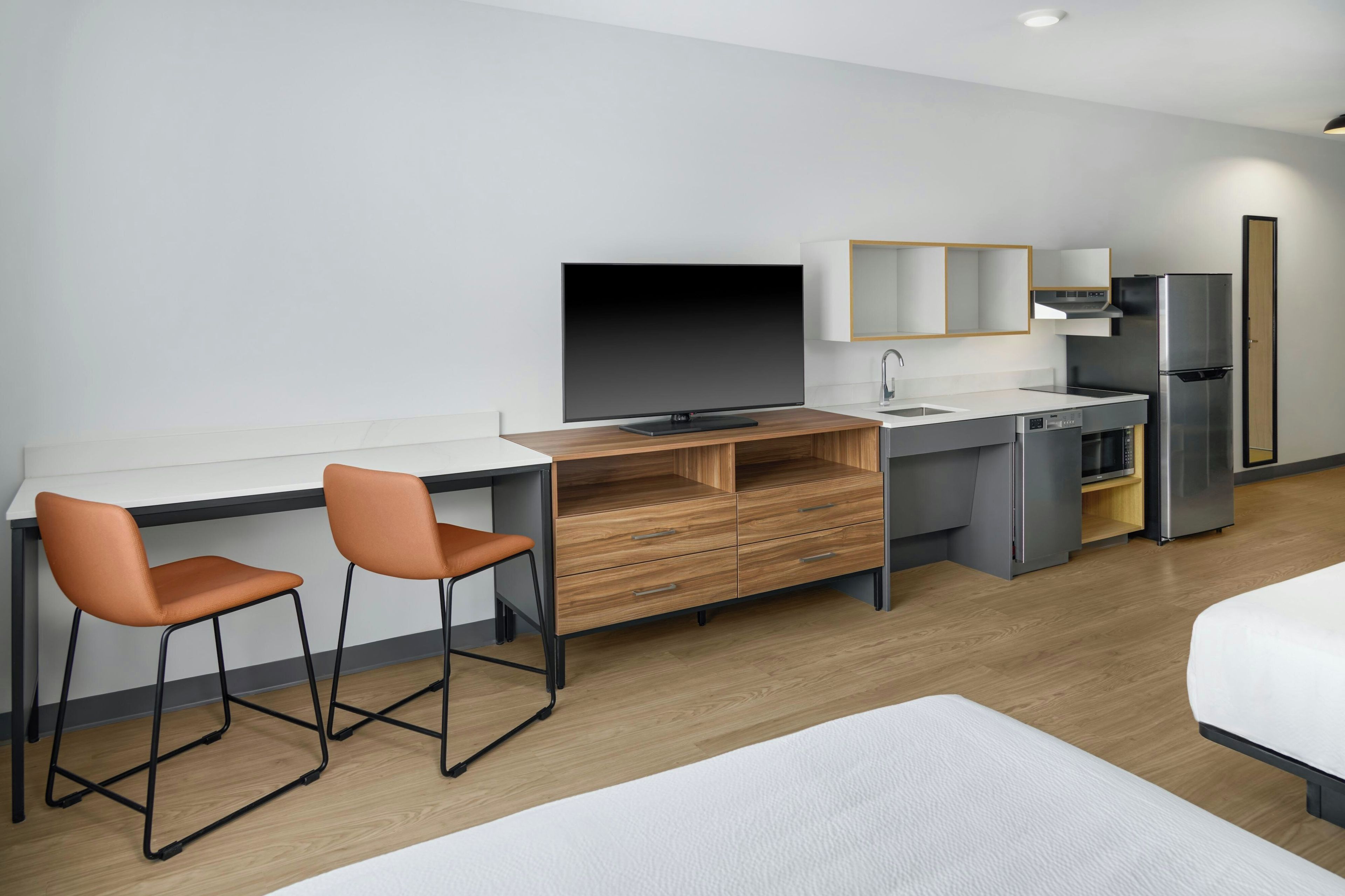 A modern hotel room with a wooden desk, two orange chairs, a flat-screen TV, a mini-fridge, a microwave, and a bed with white bedding.