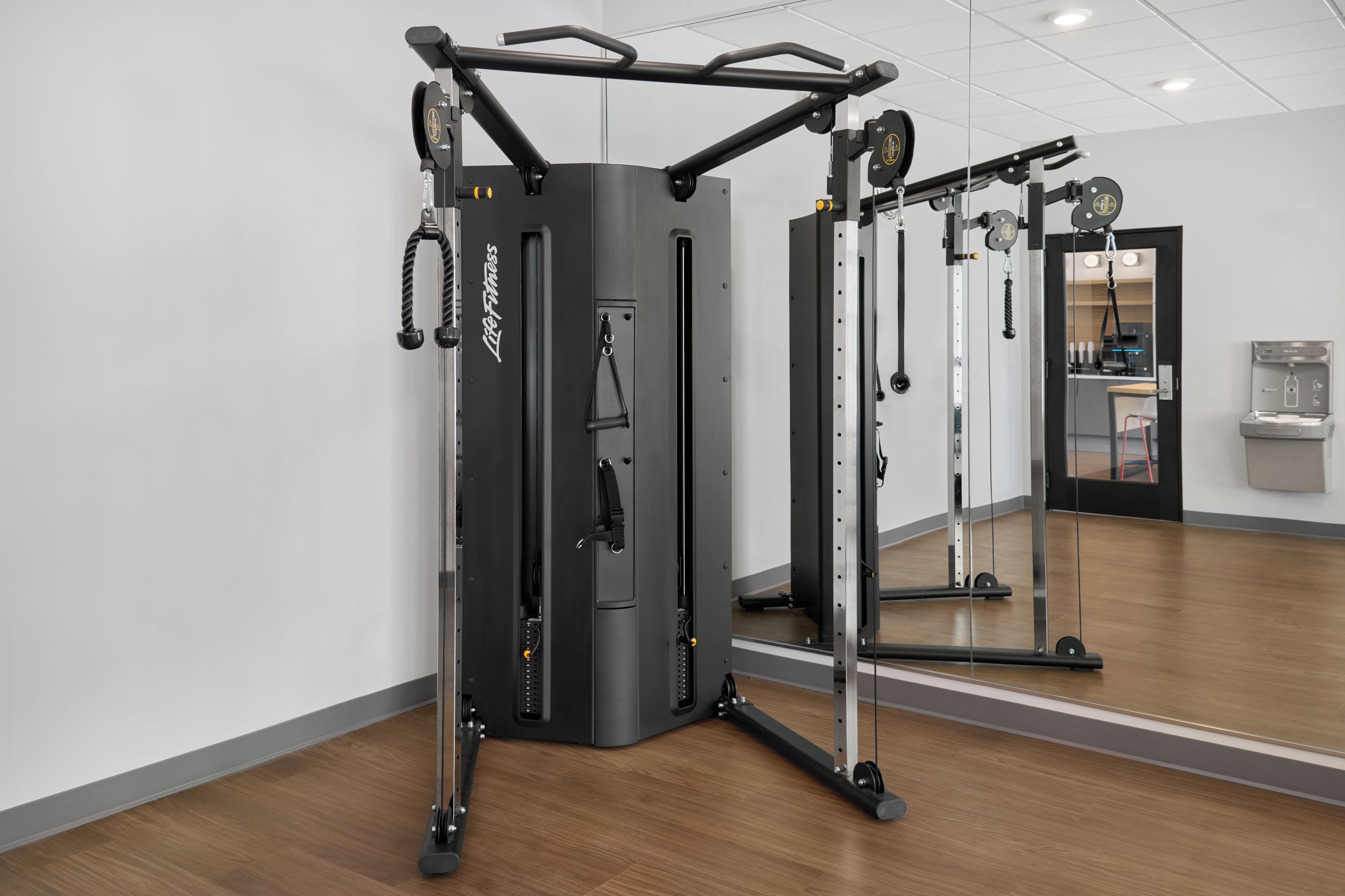 Un gimnasio con pared de espejos, equipamiento de ejercicio y dispensador de agua.