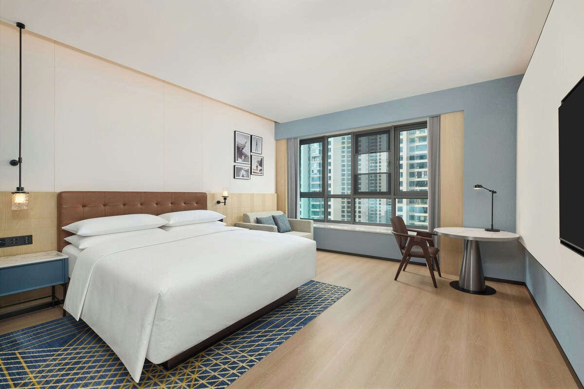 A hotel room with a bed, a desk, a chair, a window with a city view, and a wall-mounted TV.
