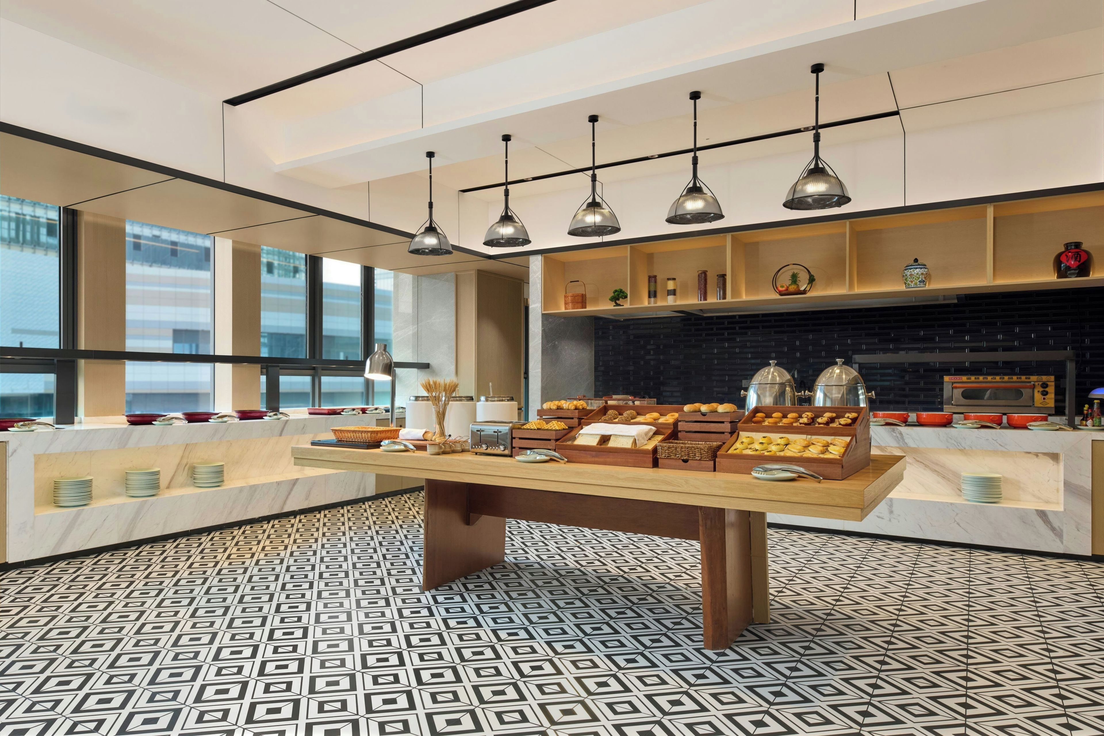 A modern kitchen with a marble countertop, a central island with food items, pendant lighting, and a view through large windows.