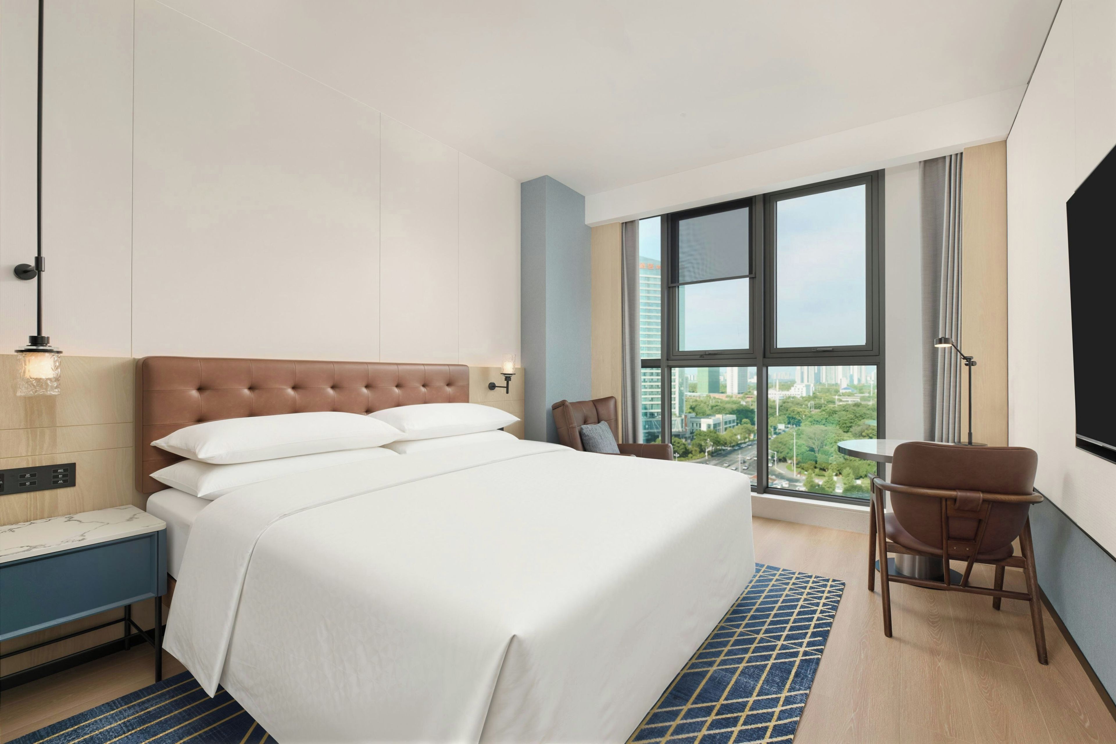 A hotel room with a large bed, a desk, a chair, and a view of the city through large windows.