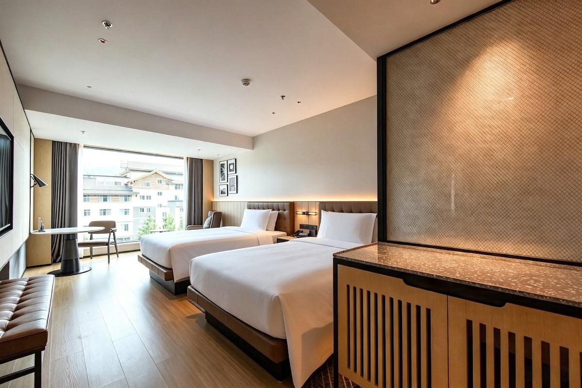 A hotel room with two beds, a desk, a chair, and a large window offering a view of buildings.