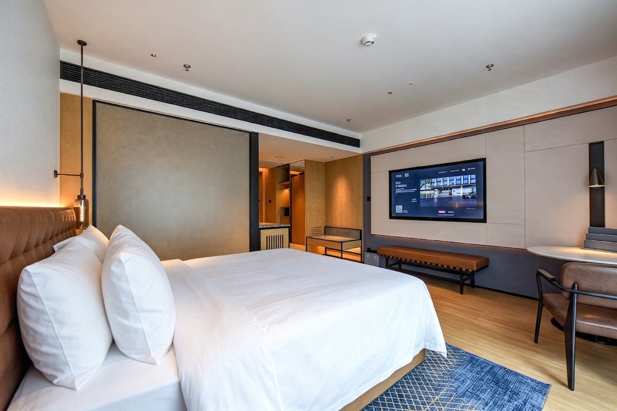 A hotel room with a large bed, a desk, a chair, a television, and a bathroom visible through an open door.