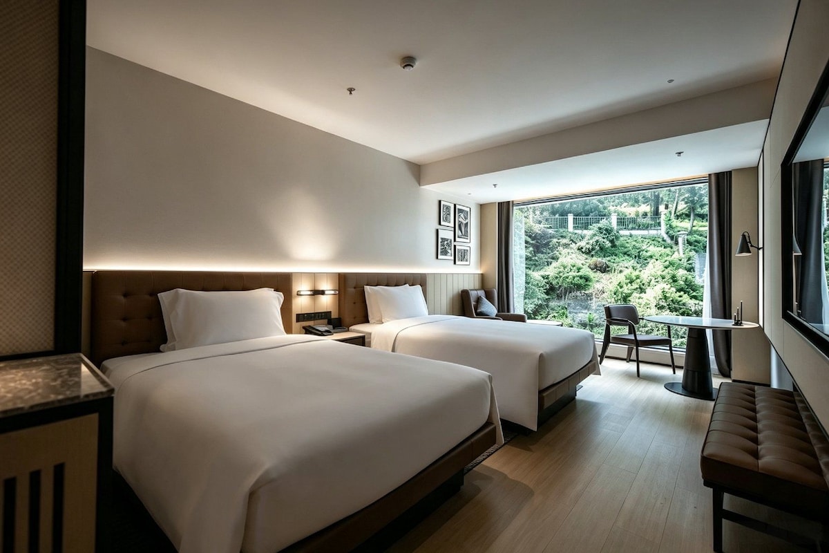 A modern hotel room with two beds, a desk, and a view of greenery outside.