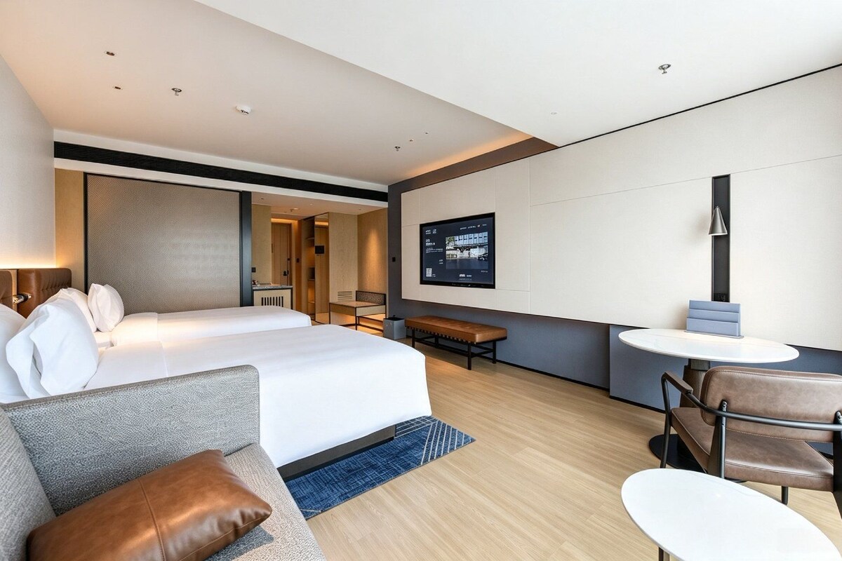 A modern hotel room with a large bed, a sofa, a desk, and a television mounted on the wall.