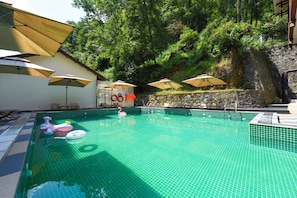 A swimming pool with a tiled floor, surrounded by lounge chairs and umbrellas, with a stone wall and greenery in the background. - Four Points By Sheraton Zhangjiajie National Forest Park (Zhangjiajie)