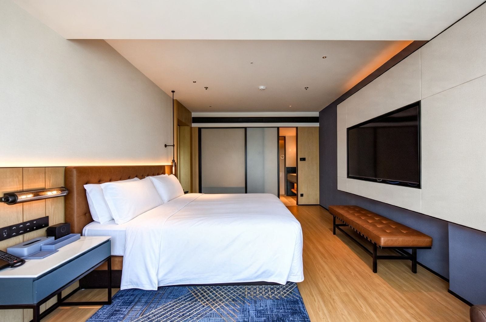 A modern hotel room with a bed, desk, bench, and a large TV.