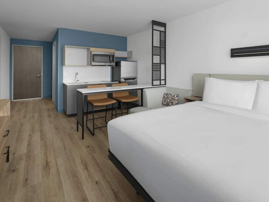 Habitación de hotel con cama, escritorio con silla, microondas, refrigerador y una pequeña zona de cocina.