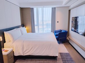 Une chambre d’hôtel avec un grand lit, un fauteuil bleu, un téléviseur fixé au mur et une vue sur les bâtiments environnants.