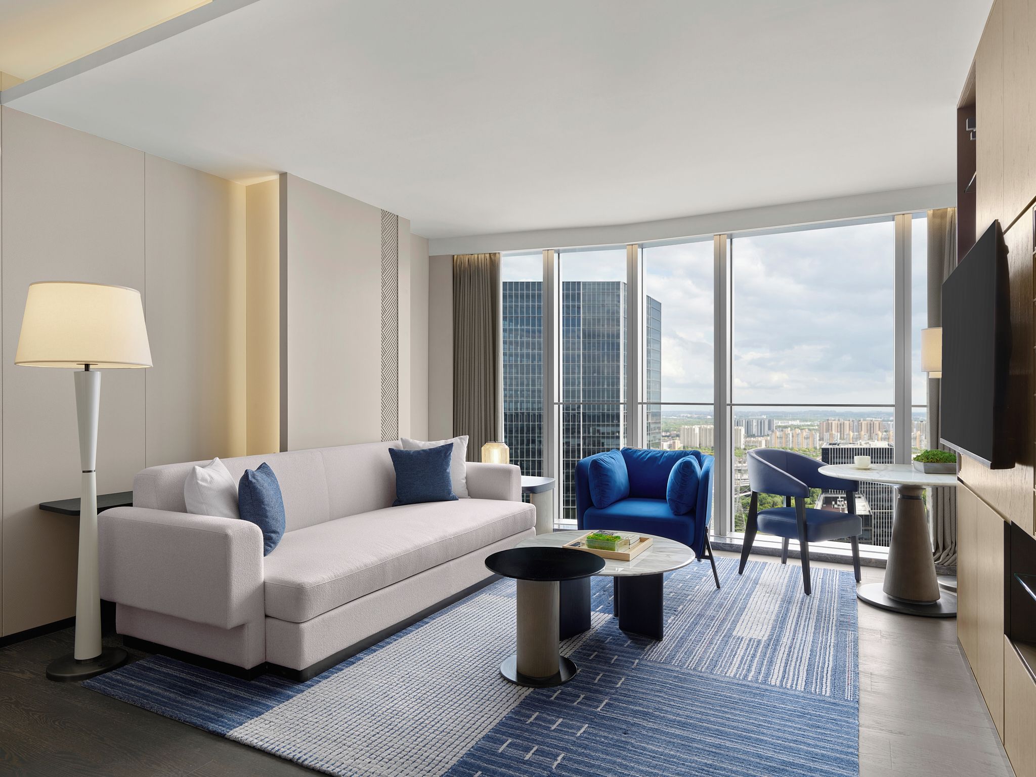 A modern hotel room with a sofa, armchair, coffee table, and a view of the cityscape through large windows.
