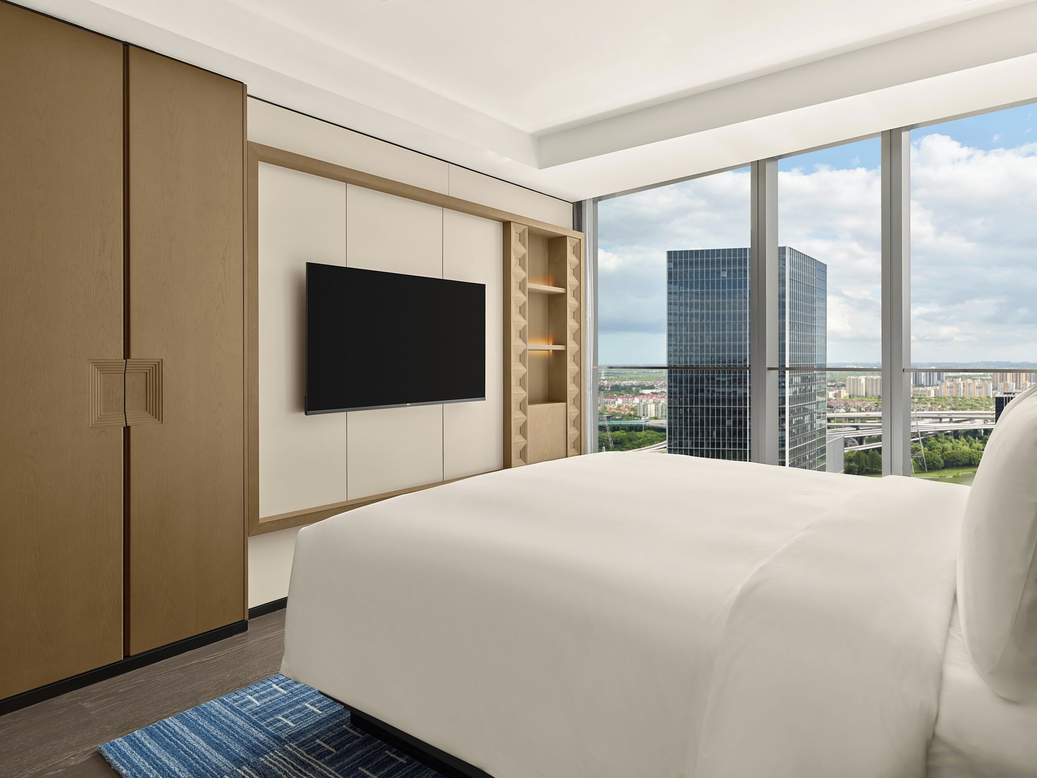Ein Hotelzimmer mit einem großen Bett, einem flachbildfernseher an der Wand und Blick auf eine Stadtlandschaft durch große Fenster.