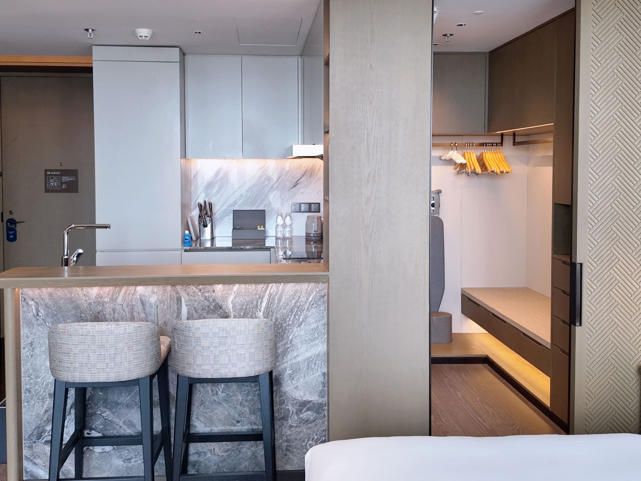 A modern hotel room with a marble bar, two bar stools, a bed, and a built-in wardrobe with hanging clothes.
