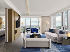 Une chambre d’hôtel moderne dotée d’une grande fenêtre, d’un canapé, d’un fauteuil, d’une table basse et d’un téléviseur fixé au mur.