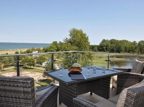 Un balcon doté de meubles en osier et d’une table en verre, offrant une vue sur une étendue d’eau et une zone herbeuse.