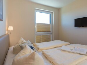 A hotel room with a bed, pillows, a bedside lamp, a window with blinds, and a flat-screen TV mounted on the wall. - Holiday Apartment With 1 Bedroom (Heiligenhafen)