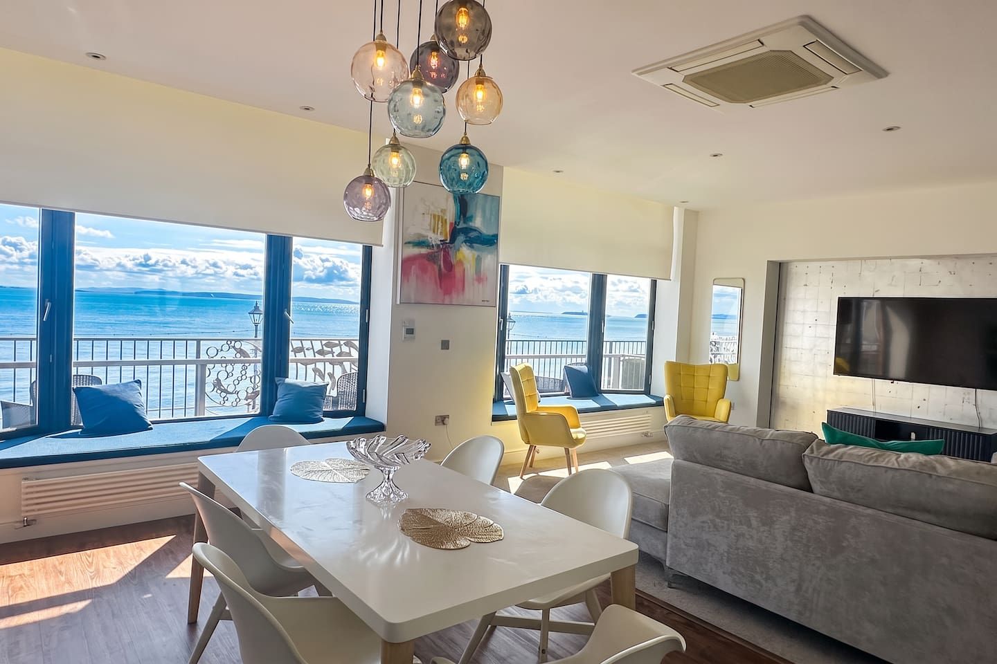 A modern living room with a large window overlooking the sea, a dining area with a white table, and a grey sofa.