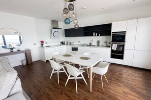 A modern kitchen with white cabinetry, a dining table with chairs, and a built-in oven. - The Hideaway | Stunning Sea Views & Stylish Stay (Vale of Glamorgan)