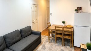 A compact living space with a grey sofa, a wooden dining table with chairs, a white refrigerator, and a door leading to another room.