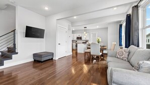 Ein modernes Wohnzimmer mit einem grauen Sofa, einem Essbereich mit Tisch und Stühlen, einer Küche mit weißen Schränken und einer Treppe, an der ein Fernseher angebracht ist.